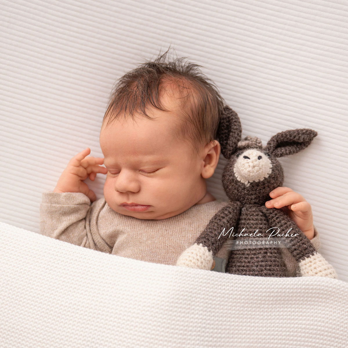 Schlafendes Neugeborenes unter einer weißen Decke, kuschelnd neben einem gehäkelten kleinen Esel.